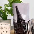 a tall, black and white photograph of the Eiffel Tower, which is a famous landmark in Paris, France. The photograph is displayed on a cylindrical container, which also has a lid and a small white ceramic container with a hole pattern. The background of the image is a white wall, and there are some green plants visible in the scene.